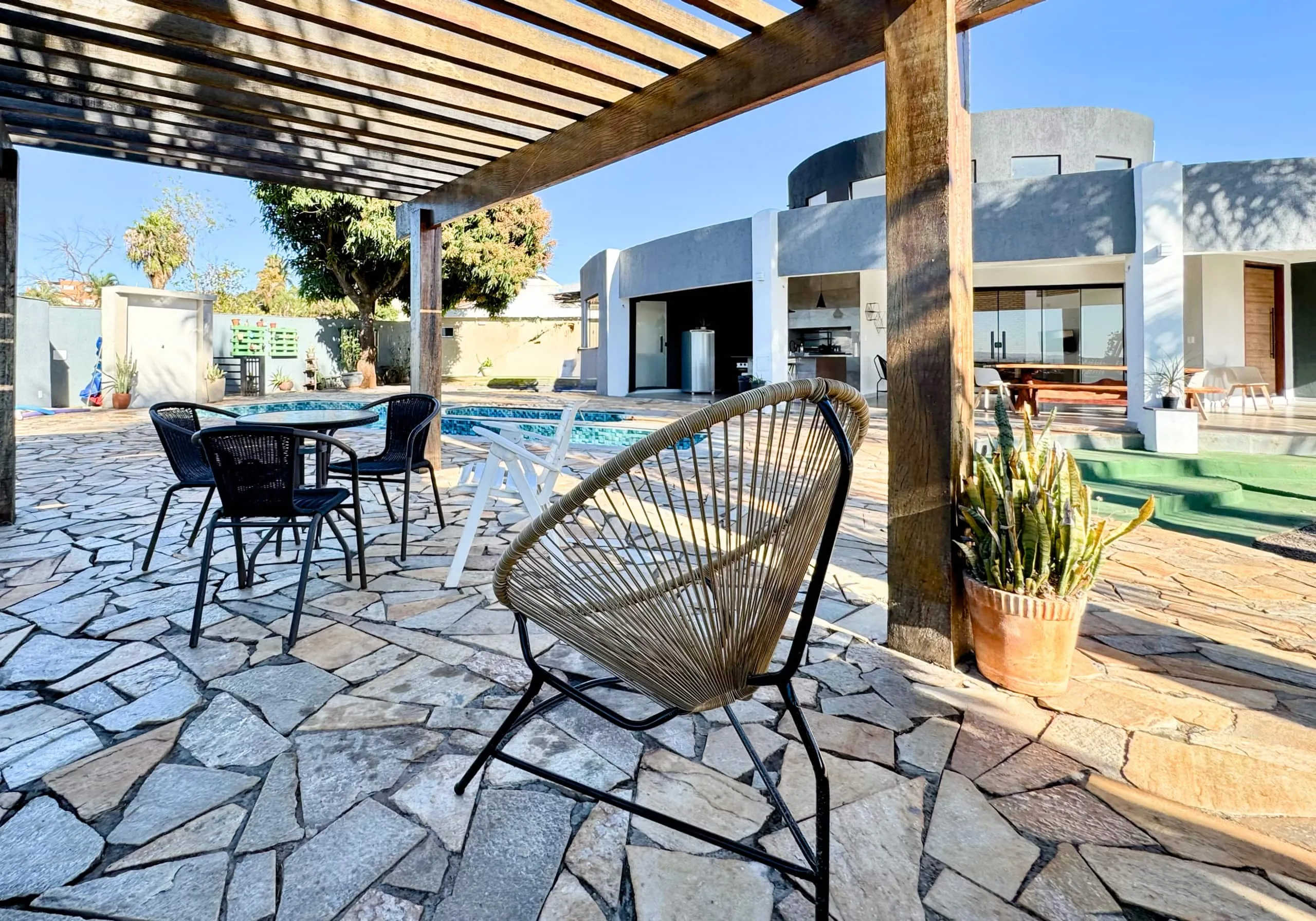 Pergolado de madeira com cadeira de fibra trançada, mesa e piscina ao lado, skyline da cidade visível ao fundo.