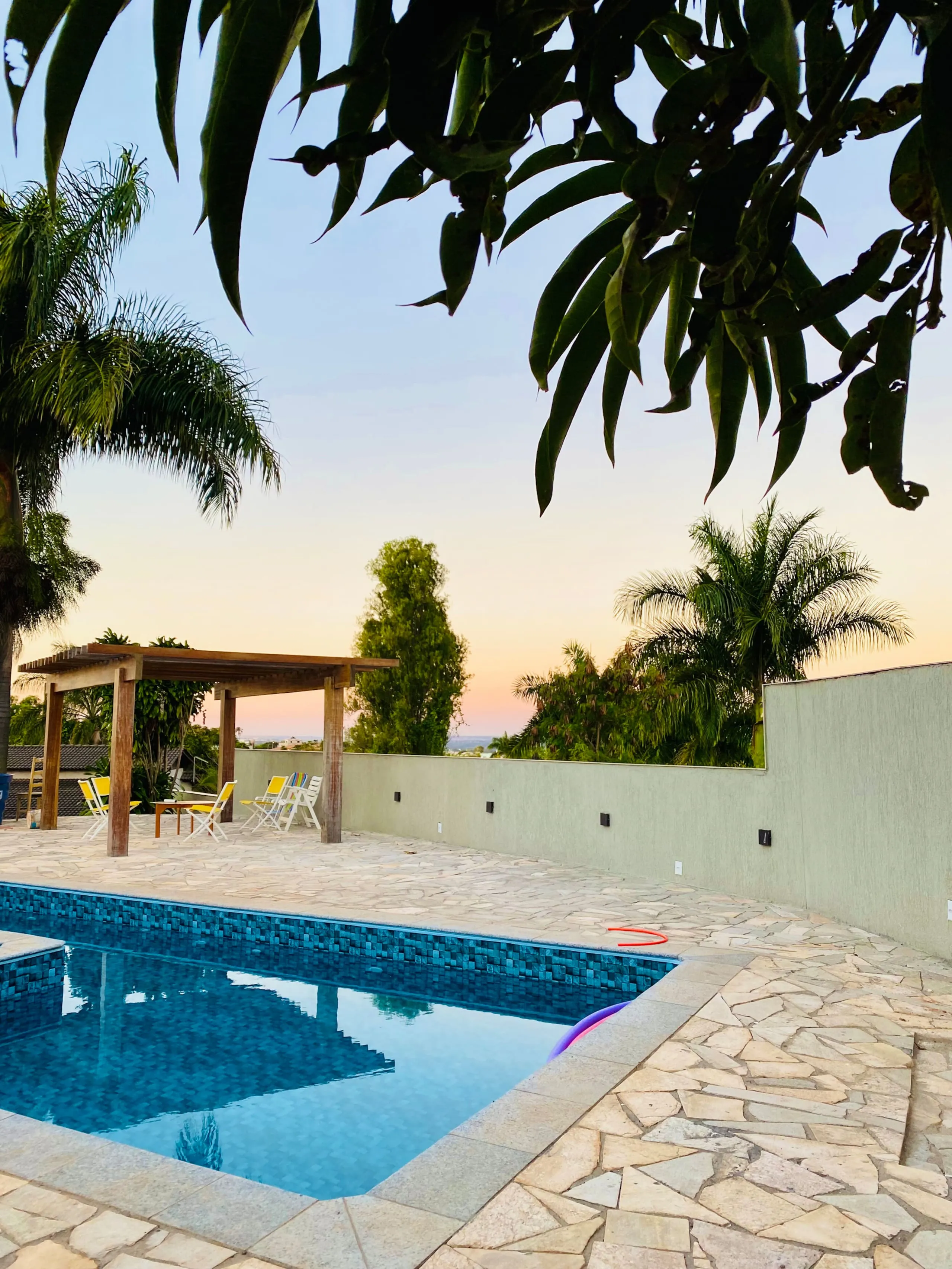 Piscina ao entardecer, com palmeiras e pergolado de madeira recortados contra o céu rosa e azul.