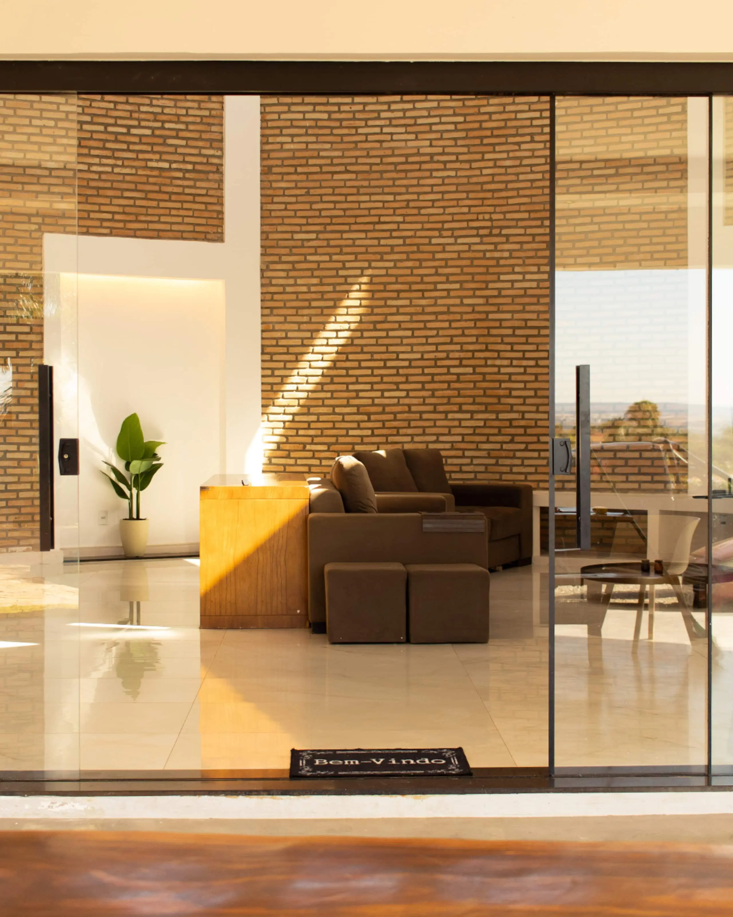 Sala vista da varanda ao fim da tarde, com a parede curva de tijolo aparente iluminada por um feixe de luz diagonal.
