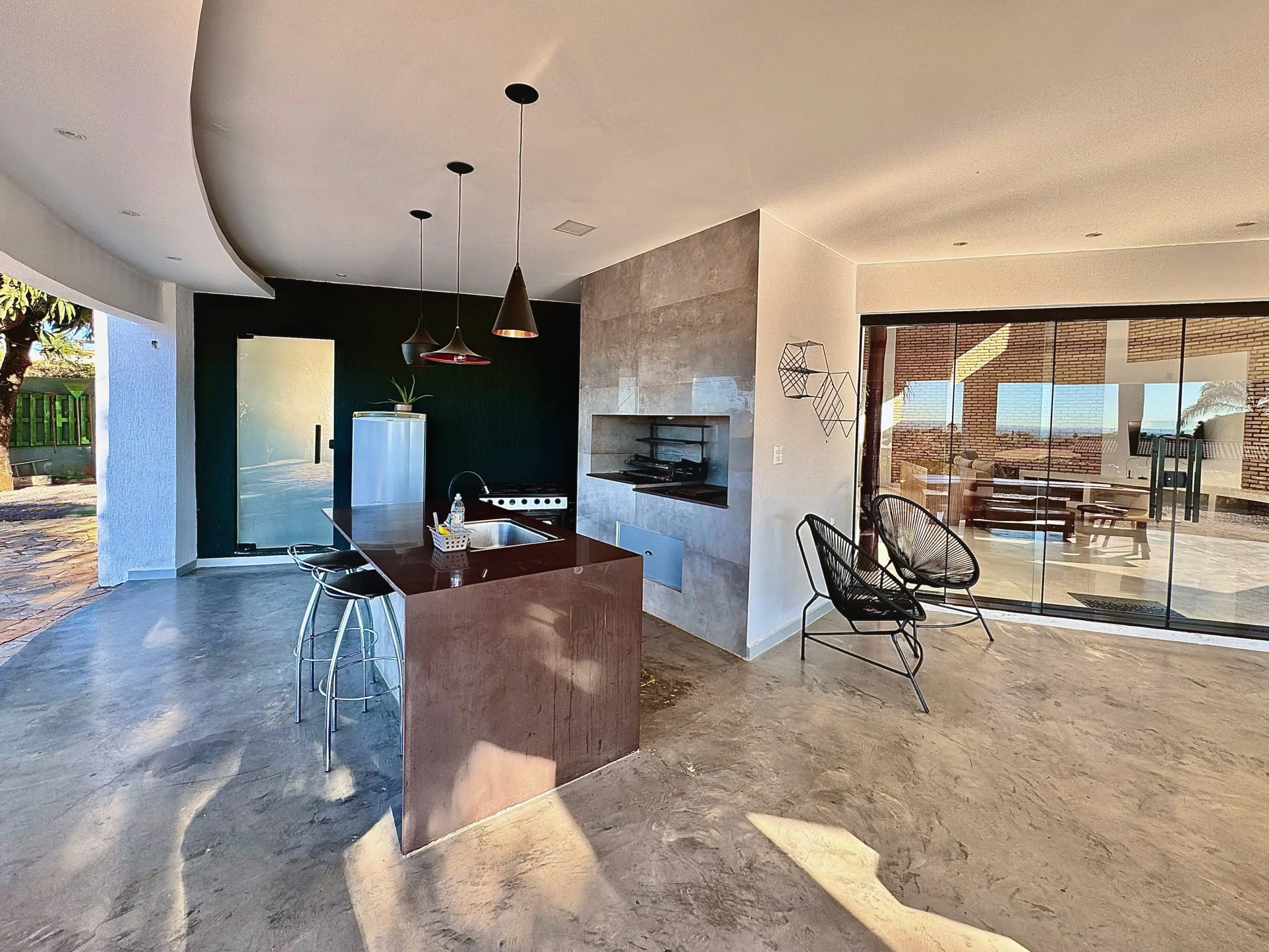 Cozinha da varanda com churrasqueira embutida, bancada e cadeiras de fio, com vista para a sala através do vidro.