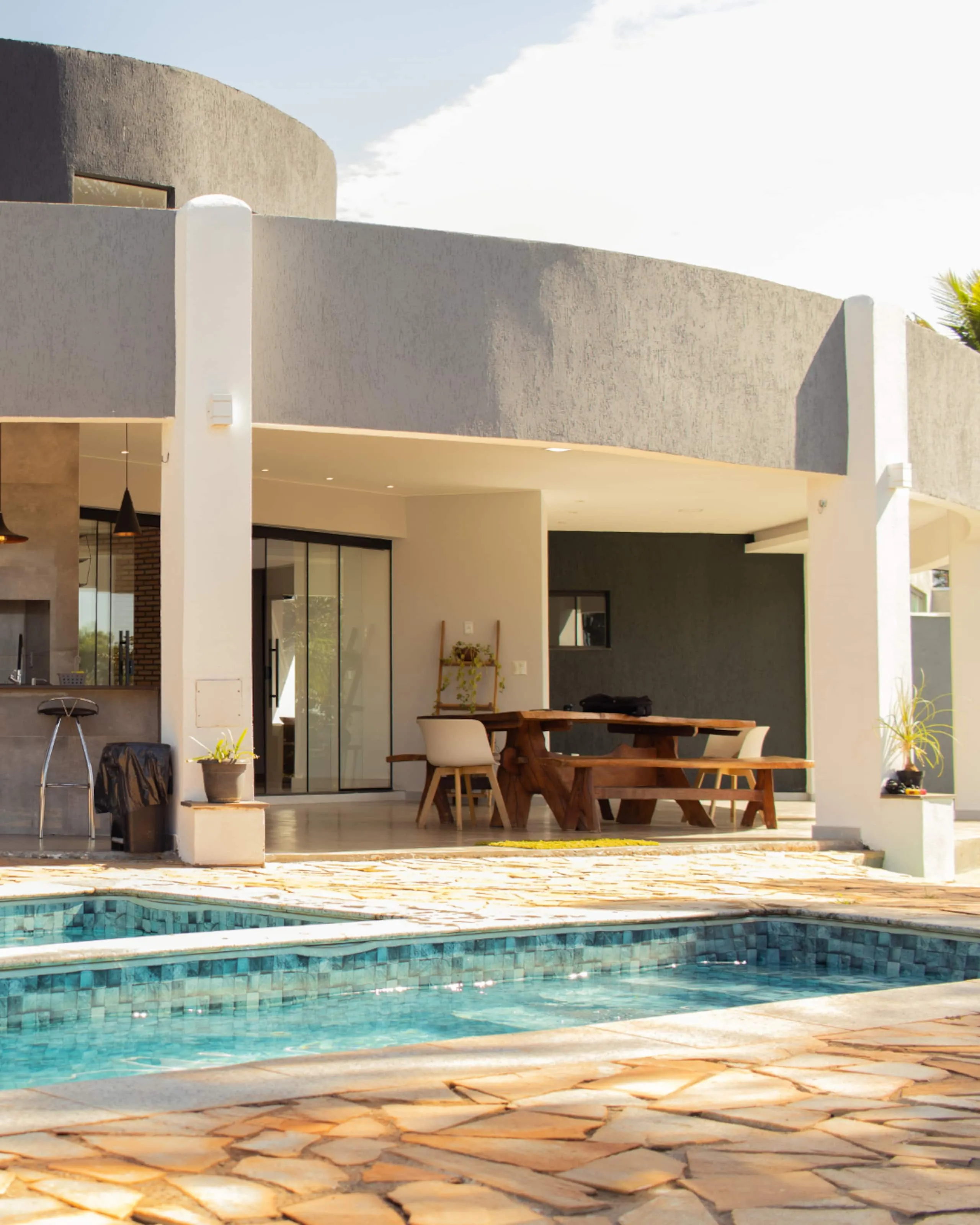 Varanda coberta com mesa de madeira maciça e piscina em primeiro plano, sob o volume curvo da casa.